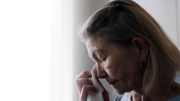 Depressziós idős, magányos nő, aki zsebkendővel törli a könnyeit, miközben az ablakon át néz. - Fotó, kép