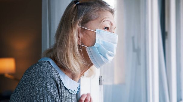 Portrait of elderly woman with medical face mask in quarantine looking through the window - Photo, Image