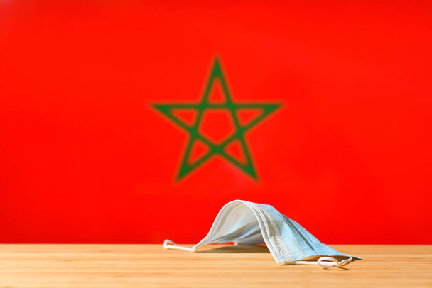 Na stole leży maska medyczna na tle flagi Maroka. Koncepcja systemu obowiązkowej maski dla mieszkańców kraju i turystów w Maroku podczas pandemii. - Zdjęcie, obraz