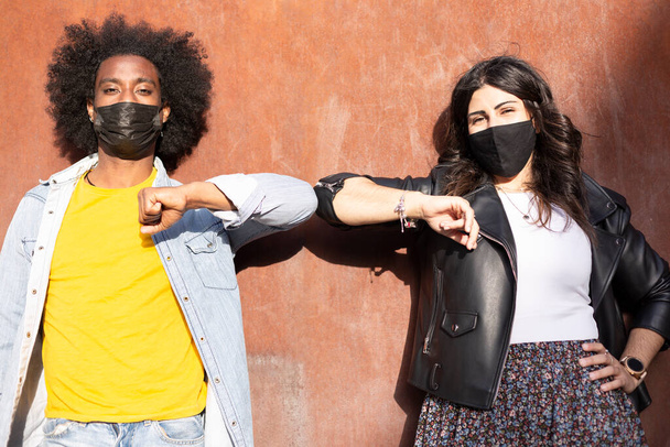 Couple of multiethnic friends wearing protective masks greet each other by bumping elbows and keeping the recommended distance to avoid Covid-19 virus contagion. - Foto, imagen