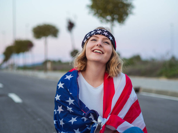 A blonde Caucasian woman from Spain sitting on a road smiling with a USA flag over her shoulders - Φωτογραφία, εικόνα