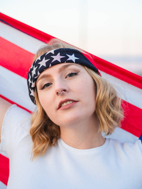 A closeup vertical shot of a blonde Caucasian woman from Spain holding the American flag behind her - 写真・画像