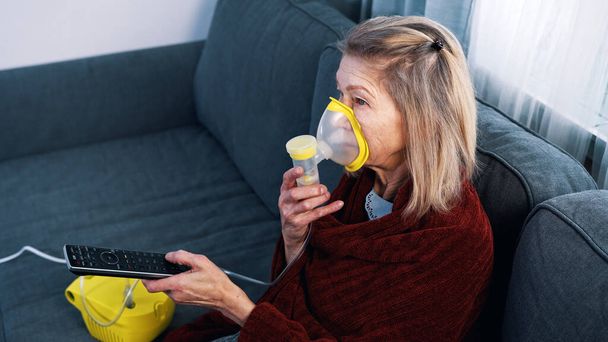 Vulnerable old woman in quarantine using oxygen inhaler and changing chanels on tv - Photo, Image