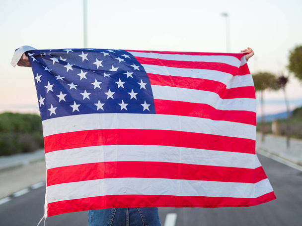A blonde Caucasian woman from Spain in jeans holding the American flag behind her - Fotó, kép