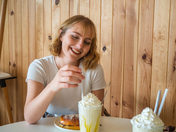 A beautiful blonde Caucasian woman from Spain sitting at a white table with two milkshakes and a donut in front of her - Fotografie, Obrázek
