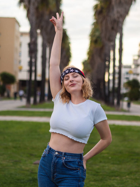 A vertical shot of a blonde Caucasian woman from Spain wearing a white crop top who has one hand raised in a peace sign and eyes closed in a park - Photo, image