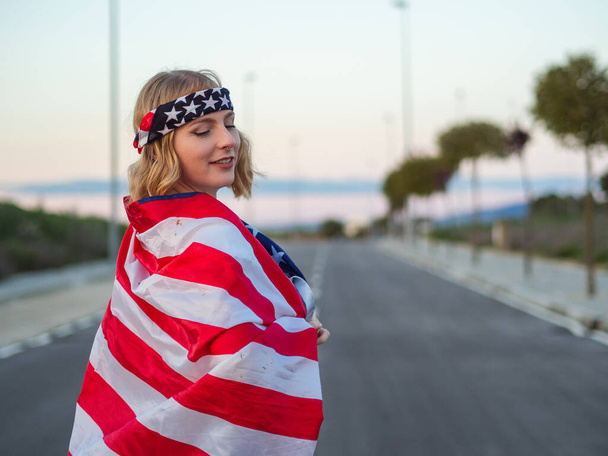 A blonde Caucasian woman from Spain covered in the American flag while wearing a USA headband - Foto, imagen