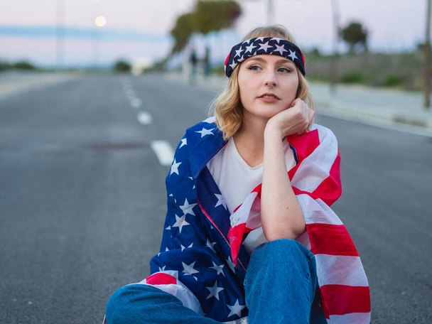 A blonde Caucasian woman from Spain sitting on a road, gazing to the side with a USA flag over her shoulders - Valokuva, kuva