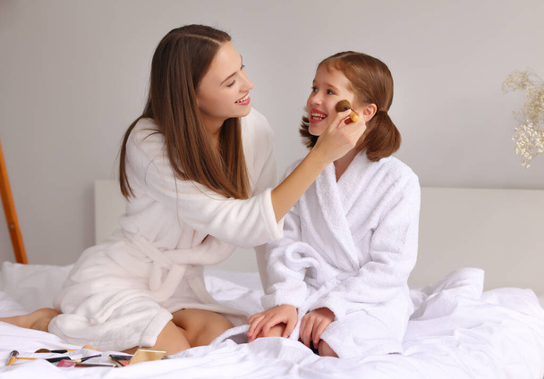 Cheerful young woman in bathrobe smiling and applying powder on cheeks of delighted girl while resting on bed during skin care routine - 写真・画像