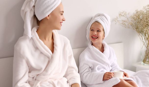 Feliz joven familia madre e hija en batas de baño y toallas sonriendo y mirándose mientras beben bebidas calientes por la mañana en la cama - Foto, Imagen
