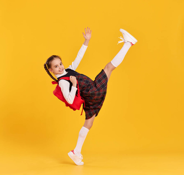 Full body side view of smiling elementary schoolgirl in checkered dress with backpack raising leg and arm while getting hurry to school against yellow background - 写真・画像