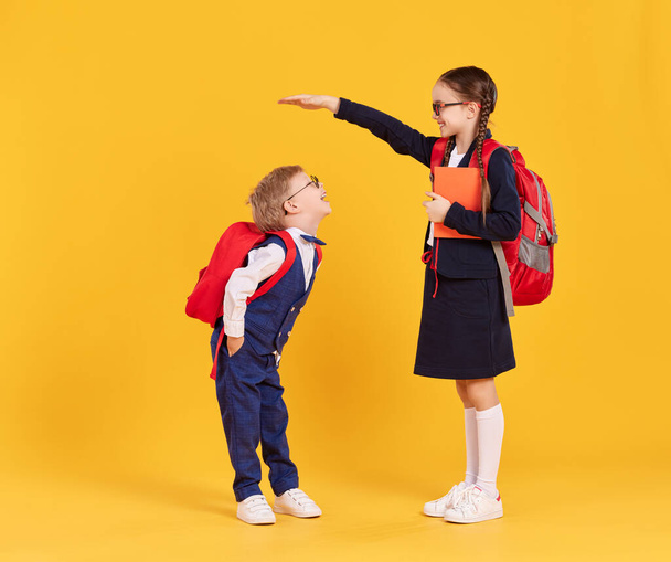 Concept of education development and growth. Happy preteen schoolgirl with backpack and book measuring height of little boy in school uniform while standing together against yellow background - Фото, изображение