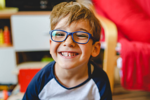 Porträt eines glücklichen kaukasischen Jungen mit Brille, der zu Hause im Zimmer sitzt und tagsüber echte Menschen kleine männliche verspielte Kinder sieht, die lächelnd in die Kamera schauen - Foto, Bild