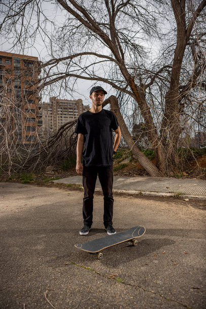 Young caucasian male in a black shirt and cap standing by his skateboard - Foto, imagen