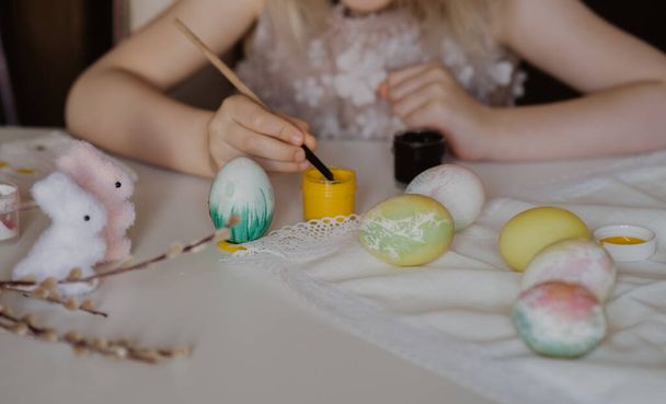 easter children's creativity. a little girl paints a white egg. near other eggs and rabbits - Fotó, kép