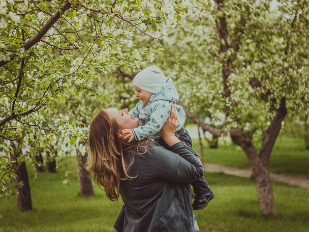 Small cute baby with his mother walking in spring park outdoor. Mom raises her little son on her hands. Blooming trees in the garden. Smile and parent care - Photo, image