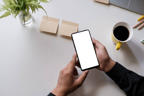 Primo piano di un uomo che tiene smartphone con schermo bianco mobile su scrivania in legno in ufficio a casa, vista dall'alto - Foto, immagini