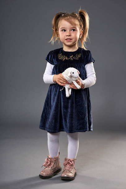 Adorable little blond girl with pigtails in deep blue dress and pink boots holding a fluffy sheep toy - Фото, изображение