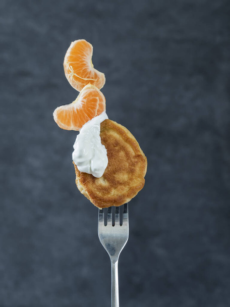 La fourchette équilibre magnifiquement les crêpes enfilées avec de la crème sure et une tranche de mandarine. Equilibre. Équilibre alimentaire. Vue latérale. Fond gris foncé. - Photo, image