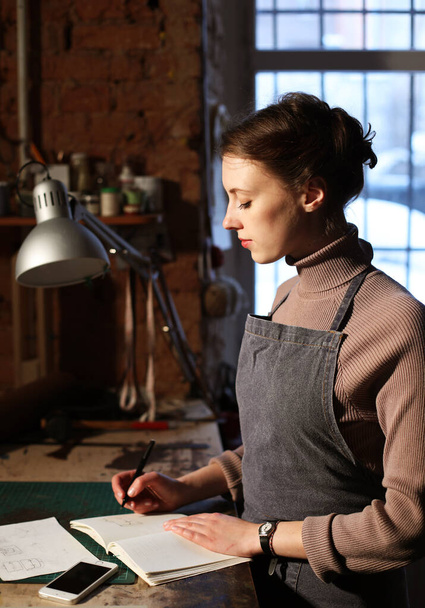 young woman writes to notebook in sewing workshop, handmade concept - Fotografie, Obrázek