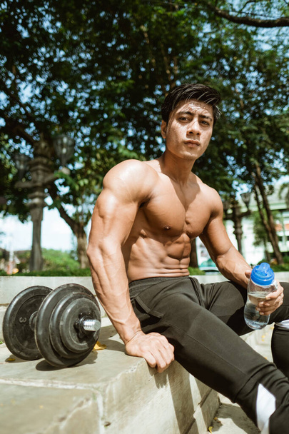 The shirtless muscular man sits holding a drinking bottle by the dumbbells after a hand muscle workout - Фото, изображение