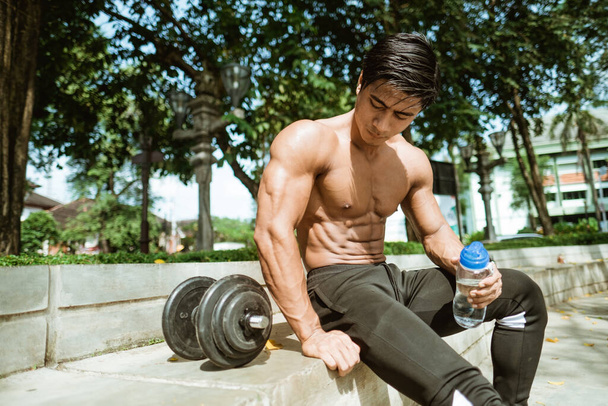 Sexy muscular man sitting poses holding a drinking bottle near dumbbells - Foto, afbeelding