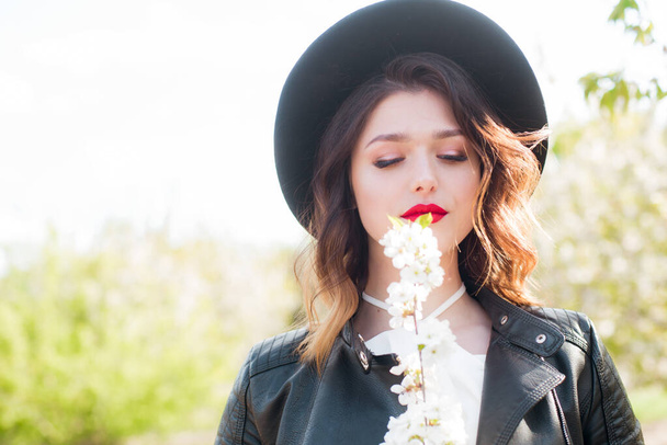 Lovely young girl in a black hat and a fashionable black leather jacket in spring in a blooming cherry garden in sunny weather with a blooming cherry branch in her hands. Spring and Fashion - Photo, image