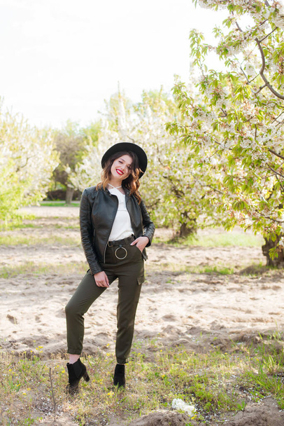 Lovely young girl in a black hat and a fashionable black leather jacket in spring in a blooming cherry garden in sunny weather with a blooming cherry branch in her hands. Spring and Fashion - Foto, Imagen