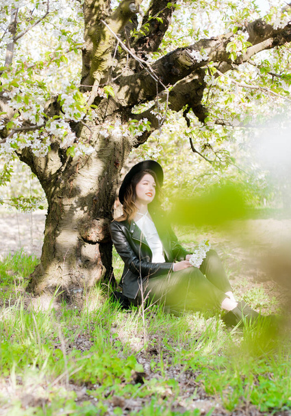 Lovely young girl in a black hat and a fashionable black leather jacket in spring in a blooming cherry garden in sunny weather with a blooming cherry branch in her hands. Spring and Fashion - Fotó, kép