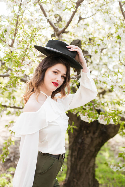 Piękna młoda dziewczyna w czarnym kapeluszu i w białej romantycznej jedwabnej bluzce wiosną w kwitnącym wiśniowym ogrodzie w słoneczną pogodę z kwitnącą gałązką wiśni w rękach. Wiosna i moda - Zdjęcie, obraz