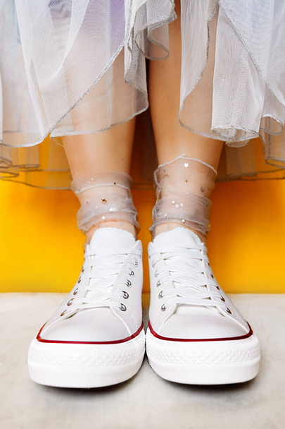 women legs in white clean new sneakers, transparent thin socks with silver shiny stars and lush tulle dress on yellow background. combination of sports shoes and formal wear. selective focus - Photo, Image