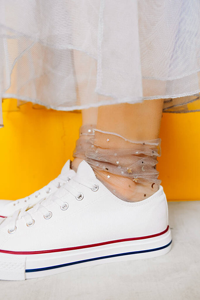 naisten jalat valkoinen puhdas uusi lenkkarit, läpinäkyvä ohut sukat hopea kiiltävä tähteä ja rehevä tylli mekko keltaisella pohjalla. yhdistelmä urheilujalkineet ja muodollinen kulumista. valikoiva kohdentaminen - Valokuva, kuva