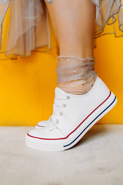 naisten jalat valkoinen puhdas uusi lenkkarit, läpinäkyvä ohut sukat hopea kiiltävä tähteä ja rehevä tylli mekko keltaisella pohjalla. yhdistelmä urheilujalkineet ja muodollinen kulumista. valikoiva kohdentaminen - Valokuva, kuva