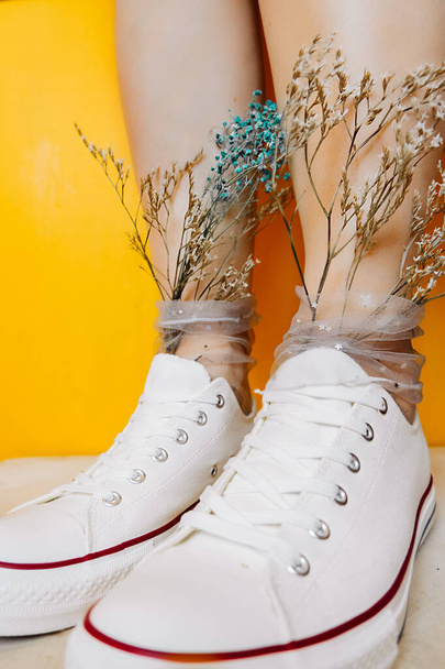 women legs in white clean new sneakers, transparent thin socks with silver shiny stars and and dry flowers in them on yellow background. combination of sports shoes and formal wear. selective focus - Photo, Image