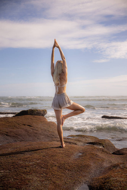Retiro de yoga en Bali. Mujer delgada practicando Vrikshasana, Tree Pose en la playa. De pie sobre la piedra. Manos levantadas en namaste mudra. Balance y concentración. Concepto de salud. Vista desde atrás. - Foto, Imagen