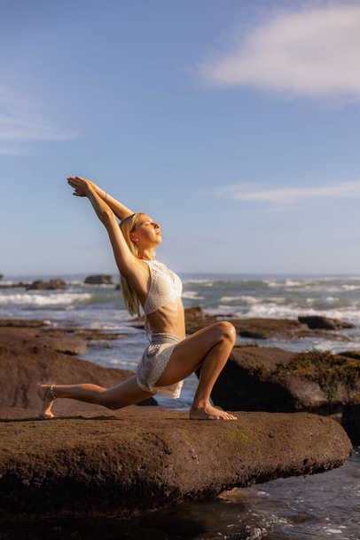 Bali yoga de playa. Mujer delgada practicando Ashwa Sanchalanasana, Pose Ecuestre. Cuerpo en forma. Estilo de vida saludable. Concepto de autocuidado. Vista al mar. Cielo azul. Copiar espacio. Bali, Indonesia. - Foto, imagen
