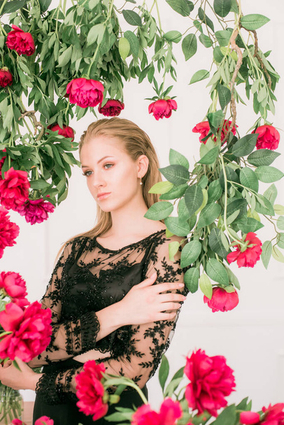 An attractive young girl with long blond hair and evening make-up in a black long evening dress surrounded by live burgundy peonies. Spring mood. Beauty and fashion  - Photo, image