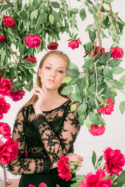 An attractive young girl with long blond hair and evening make-up in a black long evening dress surrounded by live burgundy peonies. Spring mood. Beauty and fashion  - Foto, afbeelding