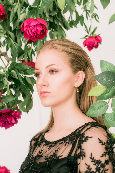 An attractive young girl with long blond hair and evening make-up in a black long evening dress surrounded by live burgundy peonies. Spring mood. Beauty and fashion  - Foto, Imagen