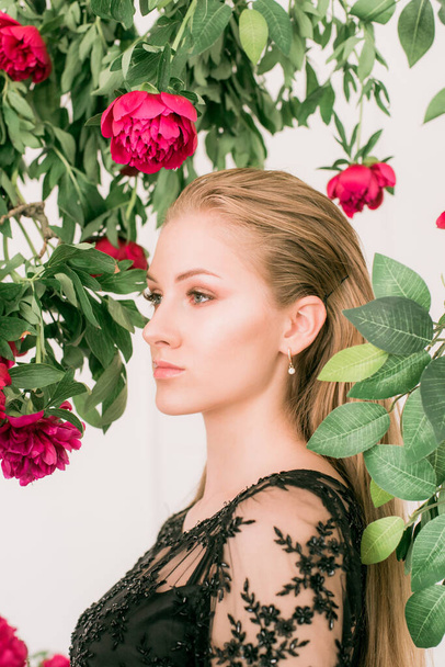 An attractive young girl with long blond hair and evening make-up in a black long evening dress surrounded by live burgundy peonies. Spring mood. Beauty and fashion  - Foto, Bild