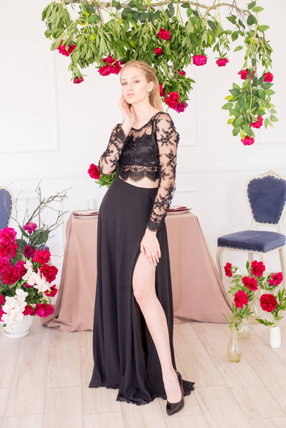 An attractive young girl with long blond hair and evening make-up in a black long evening dress, surrounded by live burgundy peonies and a festively set table for dinner. Spring mood.  - Photo, Image
