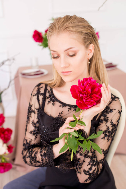 An attractive young girl with long blond hair and evening make-up in a black long evening dress, surrounded by live burgundy peonies and a festively set table for dinner. Spring mood.  - Fotoğraf, Görsel