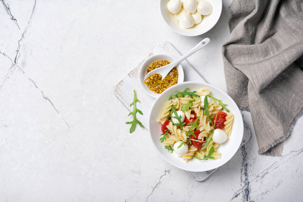 Top view of tasty vegetarian pasta with cherry tomatoes, soft cheese mozzarella, mustard sauce and green arugula leaves on white marble background - Foto, Imagem