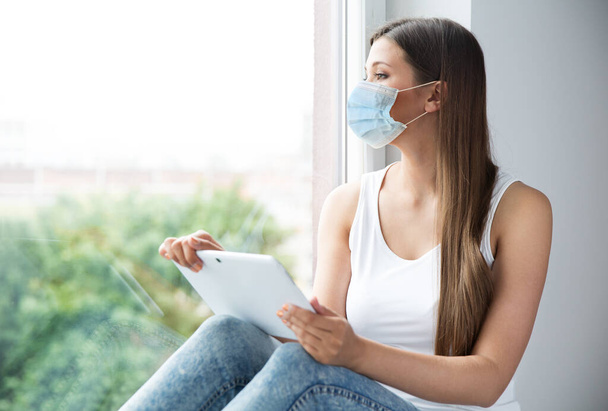 Woman in Medical Mask isolated Stay Home. Girl in Facemask Use Tablet Working at Window Sill. Self Isolation and Distance Study - Foto, immagini