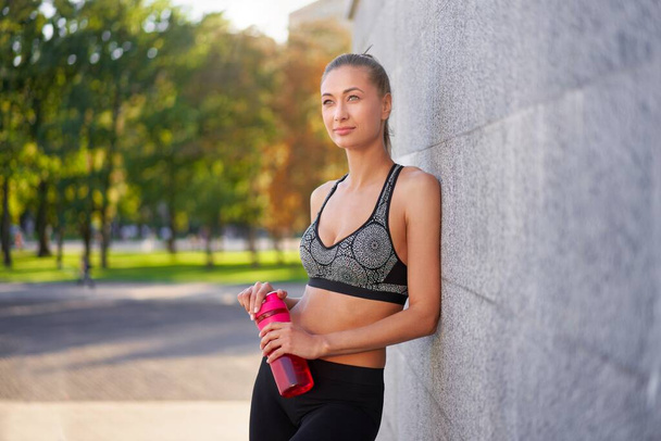 Tired Woman drink water after morning workout Young athletic female standing leaning wall city street park background after jogging Healthy lifestyle concept Copy space - Фото, зображення