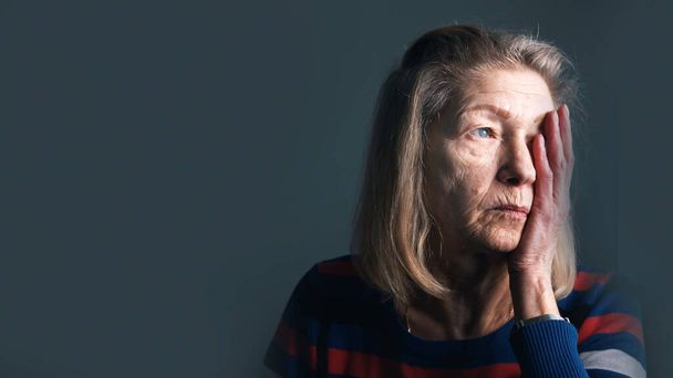 Беспокойная пожилая женщина, держащая за руку лицо и глядя в окно. Одиночество или проблемы с глазами - Фото, изображение
