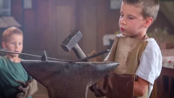 Niño pequeño en taller de forja de metal usando martillo para batir la barra de metal - Imágenes, Vídeo