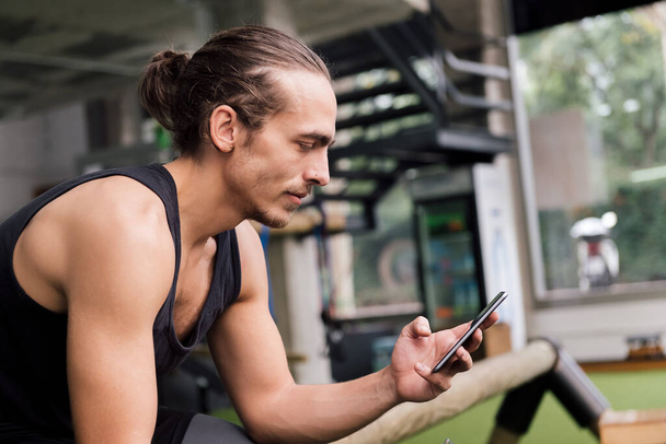 young athlete consulting the phone while resting from his workout at the gym, concept of technology and active lifestyle - Foto, Bild