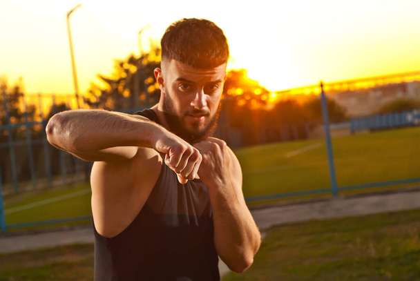Athlete trains on a sunset background. The boxer warms up near the stadium. Contrasting dramatic shadows on the face as an artistic effect. The face is blurred, the sharpness on the fist. - Фото, изображение