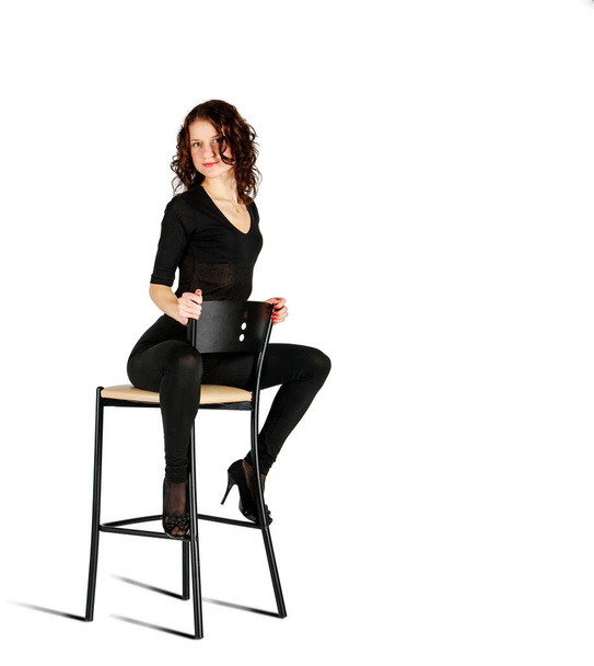 Siyah takım elbiseli genç kız, beyaz arka planda, stüdyoda bir sandalyede otururken poz veriyor. - Fotoğraf, Görsel